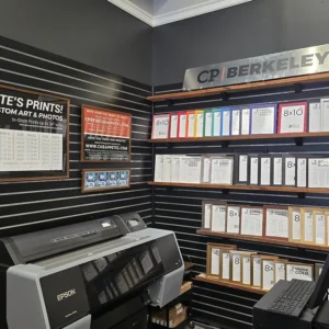 Corner of the Cheap Pete’s Berkeley store with a large Epson photo printer below a wall of black slatwall. Signs advertise in-store prints up to 24 inches wide, with a price chart. Shelves under a “CP Berkeley” sign hold ready-made frames in colored, white, gray, wood and gold finishes.